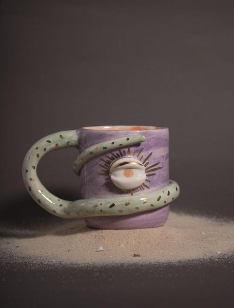 purple colour mug. there is an eye on it and its holder snake figure and wrapping around the cup on dessert sand